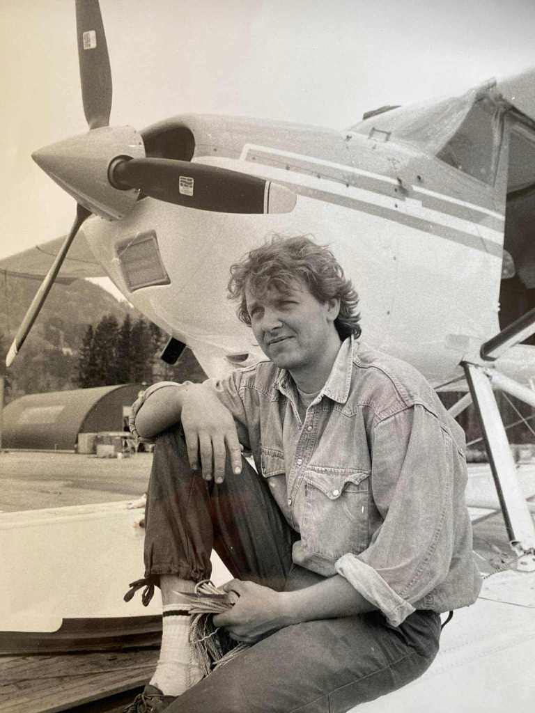 A man sitting on the wing of a small airplane, wearing a denim jacket and relaxed clothing, with a propeller and hangars in the background.