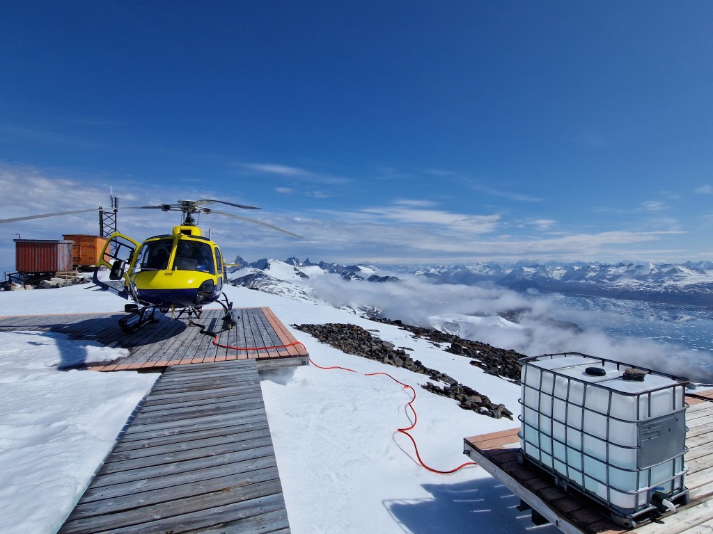 A yellow and blue helicopter parked on a wooden platform in a snowy mountainous landscape under a clear blue sky.